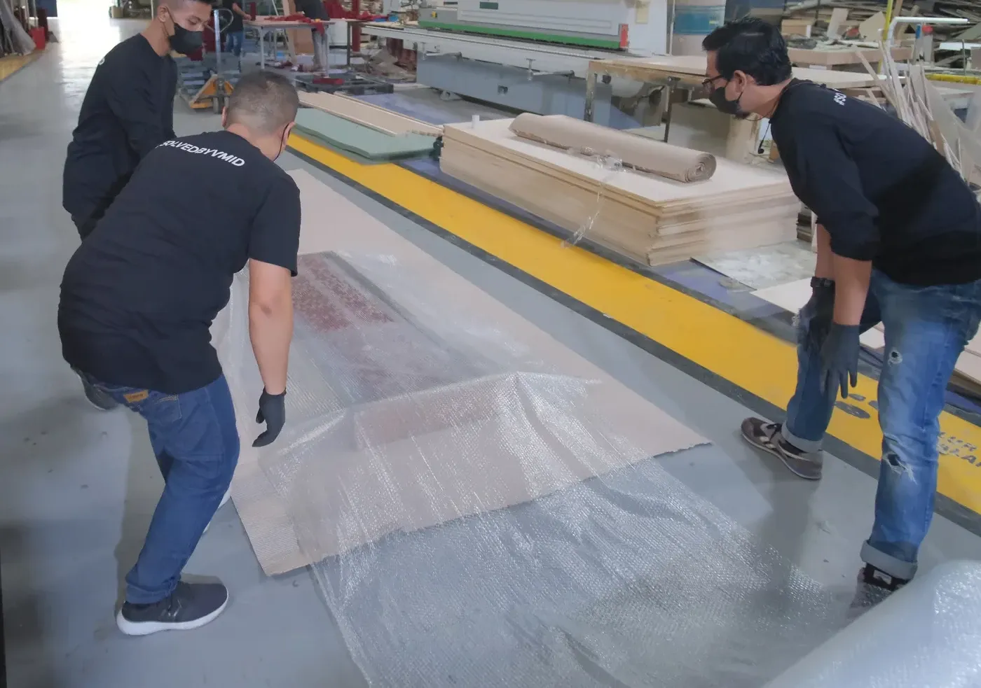 Three crew members in VMID shirts spreading plastic wrap over a panel on the workshop floor