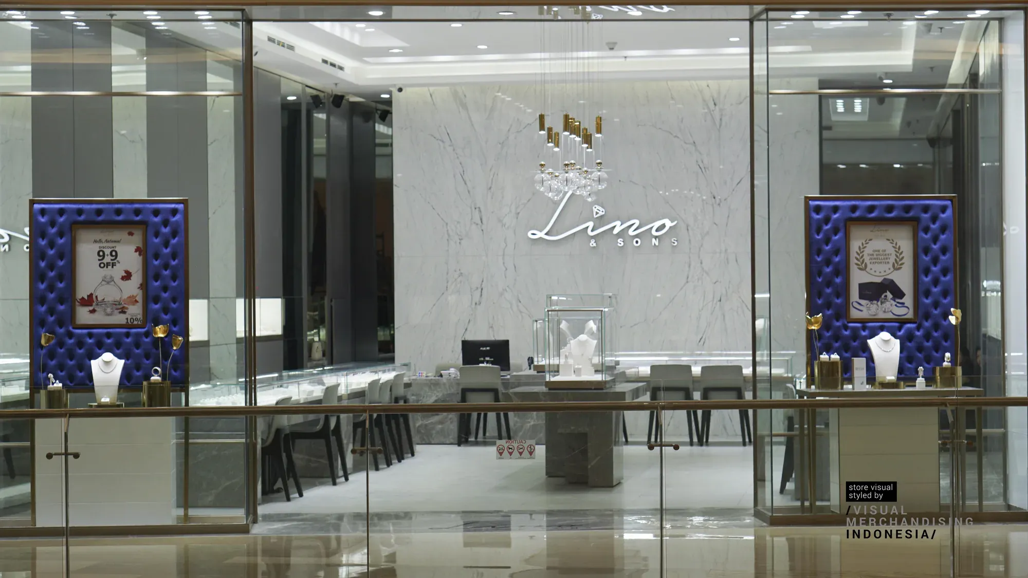 A glass shopfront opens onto a marble-lined jewellery boutique, flanked by two tufted navy-velvet display panels each holding a jewellery bust and a small framed card, with the illuminated 'Lino & Sons' sign and a cluster of pendant lights centred on the back wall