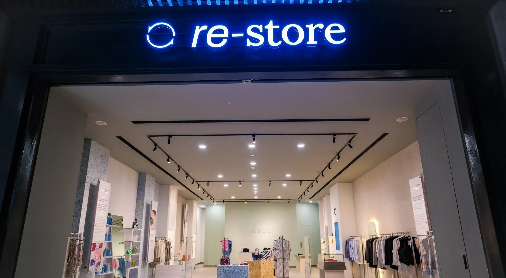 re-Store at Astha, Jakarta — the lit re-Store shopfront sign above a dark storefront frame, with the dressed interior and garment rails visible through the opening