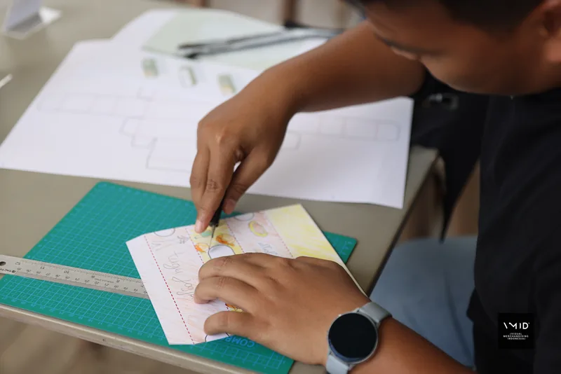 A hand drawing a coloured label onto a paper template on a cutting mat — CAMP 2023, PT. Agrinesia Raya