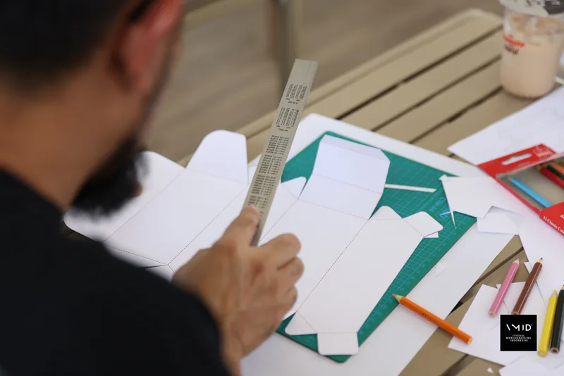 Hands folding a white paper box net against a steel ruler on a green cutting mat — CAMP 2023