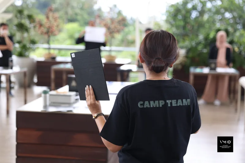 A CAMP TEAM member holding up a board in front of the tables in the open-air pavilion — PT. Agrinesia Raya, 2023