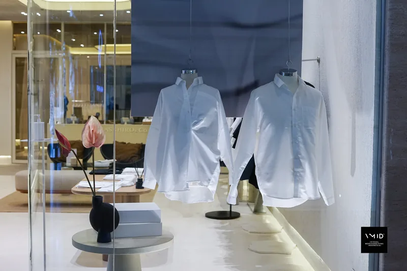 The Masshiro&Co. window from inside: two white shirts hung against a slate panel, an anthurium in a black vase on a white plinth — 2022