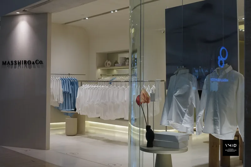 Seen from the corridor, two white shirts hung in the Masshiro&Co. window beside an anthurium in a black vase, blue and white shirts on rails inside — 2022