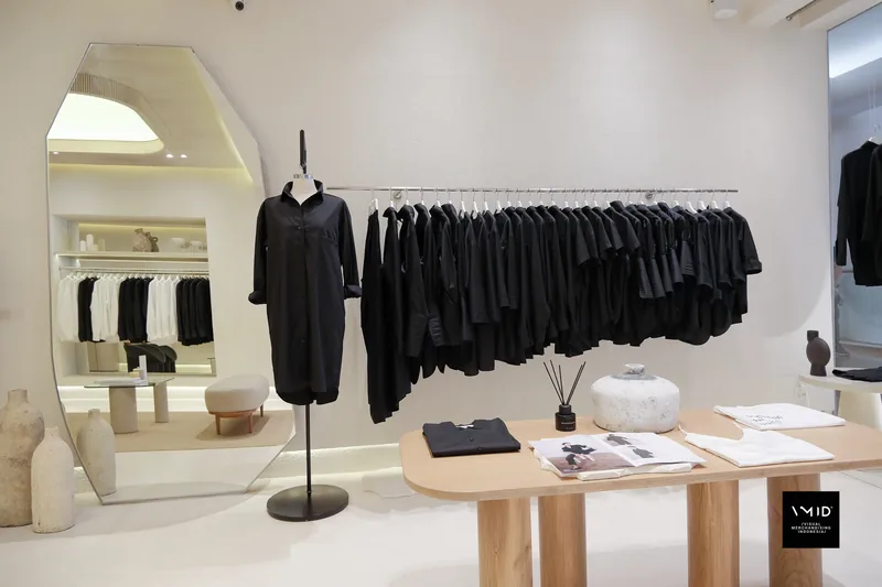 A mannequin in a black shirt dress beside a wall rail of black pieces, folded tees and a stone vase on the timber table — Masshiro&Co., 2022